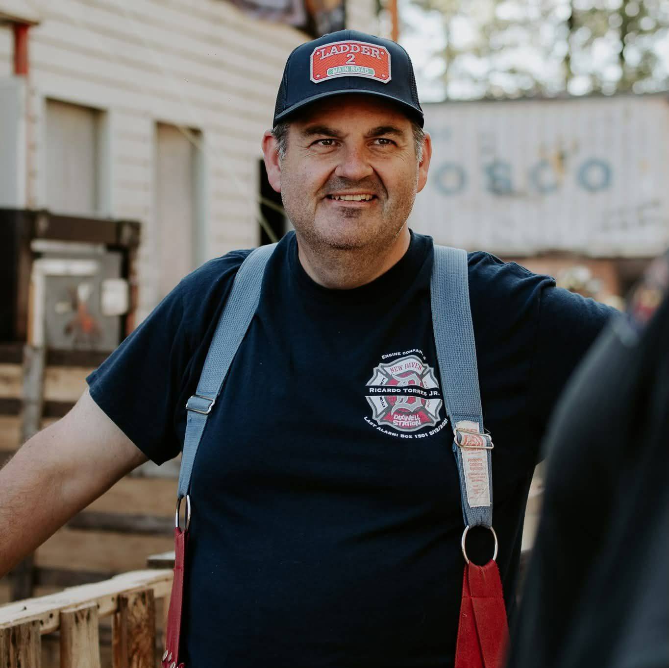 Joe Bourquin outdoors in a navy fire department cap and turnout suspenders, with a soft-focus building in the background.